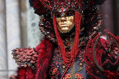 An elaborate carnival costume featuring an intricate design. The attire includes a golden mask and ornate headdress embellished with red beads, feathers, and gemstones. The costume is richly decorated, blending textures and colors to create a striking appearance.