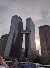 Two modern skyscrapers are connected by a bridge at an upper floor. The buildings have a sleek, glass facade and are situated against a cloudy sky with some sunlight peeking through. People, possibly tourists, are visible at the bottom, seemingly on a sightseeing excursion.