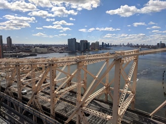 A metal bridge spans across a river with a backdrop of a city skyline. The sky is bright blue with scattered white clouds, and the cityscape features modern high-rise buildings. Vehicles can be seen on the bridge, which has intricate structural details.