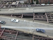 Aerial view of a multi-lane urban roadway with cars driving on two lanes. The infrastructure includes visible metal beams and supports underneath the road. Buildings and some greenery are seen in the background, indicating an urban setting.