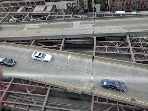 Aerial view of a multi-lane urban roadway with cars driving on two lanes. The infrastructure includes visible metal beams and supports underneath the road. Buildings and some greenery are seen in the background, indicating an urban setting.