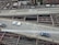 Aerial view of a multi-lane urban roadway with cars driving on two lanes. The infrastructure includes visible metal beams and supports underneath the road. Buildings and some greenery are seen in the background, indicating an urban setting.