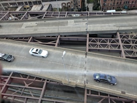 Aerial view of a multi-lane urban roadway with cars driving on two lanes. The infrastructure includes visible metal beams and supports underneath the road. Buildings and some greenery are seen in the background, indicating an urban setting.