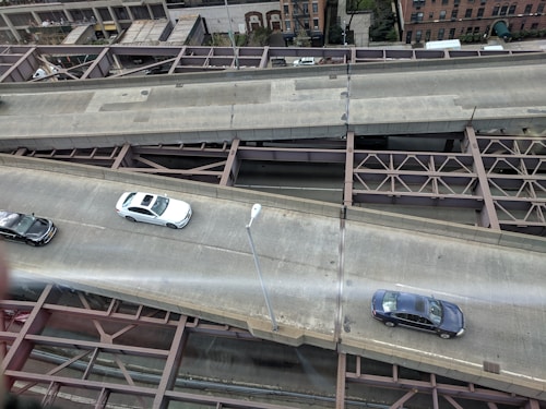 Aerial view of a multi-lane urban roadway with cars driving on two lanes. The infrastructure includes visible metal beams and supports underneath the road. Buildings and some greenery are seen in the background, indicating an urban setting.