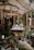 A cozy indoor garden or greenhouse setting with a variety of lush green plants and colorful flowers. Wooden structures and shelves filled with potted plants create a natural and earthy atmosphere. A central wooden counter displays items, possibly for sale, and is surrounded by hanging and standing plants. Soft light filters through a large umbrella and the glass structures, giving an inviting feel.