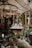 A cozy indoor garden or greenhouse setting with a variety of lush green plants and colorful flowers. Wooden structures and shelves filled with potted plants create a natural and earthy atmosphere. A central wooden counter displays items, possibly for sale, and is surrounded by hanging and standing plants. Soft light filters through a large umbrella and the glass structures, giving an inviting feel.