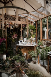 A cozy indoor garden or greenhouse setting with a variety of lush green plants and colorful flowers. Wooden structures and shelves filled with potted plants create a natural and earthy atmosphere. A central wooden counter displays items, possibly for sale, and is surrounded by hanging and standing plants. Soft light filters through a large umbrella and the glass structures, giving an inviting feel.
