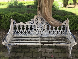 An ornate, weathered metal bench with intricate patterns, painted in fading white with patches of blue, is situated in a garden. A large tree has grown into the bench, enveloping part of it, indicating the passage of time. The bench is placed on a stone-paved ground, surrounded by neatly trimmed green bushes.