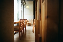 A cozy cafe interior with wooden chairs and tables arranged along a window, allowing natural light to stream in. The warm tones of the wood and soft lighting create an inviting atmosphere.