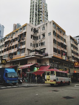 A busy urban street corner with a high-rise residential building showing signs of wear, such as chipped paint and laundry hanging from windows. The street level features a bustling scene with a van, a truck, and a motorcycle in motion. Several signs, including a fast-food restaurant and various shops, add to the urban atmosphere.