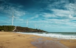 A scenic coastal landscape with several wind turbines lined up on a grassy hill near the shore. The foreground features a sandy beach with gentle waves washing up to the shoreline. The sky is expansive with scattered clouds, giving a sense of openness.