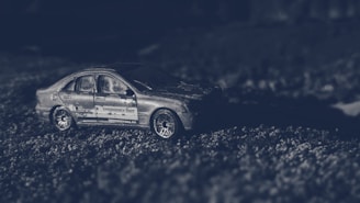 A miniature model of a car with the words 'Emergency Unit' on its side is placed on a textured surface, possibly simulating a road. The lighting is dark and atmospheric, creating a dramatic effect.