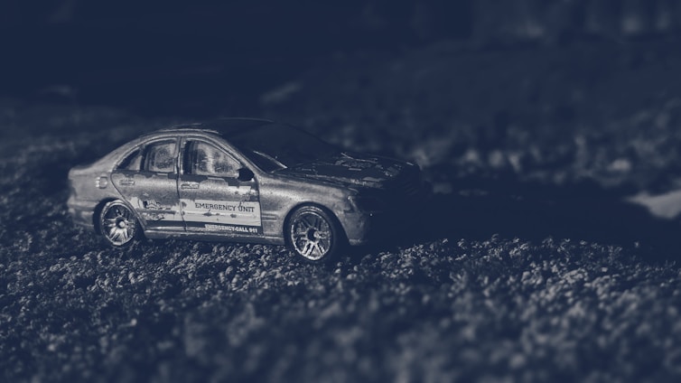 A miniature model of a car with the words 'Emergency Unit' on its side is placed on a textured surface, possibly simulating a road. The lighting is dark and atmospheric, creating a dramatic effect.
