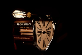 A vintage-style light bulb illuminates a stack of books, on top of which is a distorted, surreal clock reminiscent of Salvador Dali's melting clocks. The books include titles such as 'I Am Second,' 'not a fan,' and 'Fox's Book Of Martyrs.' The scene is dimly lit, creating strong contrasts between light and shadow.