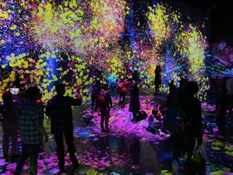 A vibrant and immersive digital art installation with bright, colorful light projections resembling flowers and abstract patterns covering the walls and floor. People are interacting with the environment, some sitting and others standing or walking around, creating an interactive and dynamic atmosphere.