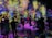 A vibrant and immersive digital art installation with bright, colorful light projections resembling flowers and abstract patterns covering the walls and floor. People are interacting with the environment, some sitting and others standing or walking around, creating an interactive and dynamic atmosphere.