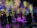 A vibrant and immersive digital art installation with bright, colorful light projections resembling flowers and abstract patterns covering the walls and floor. People are interacting with the environment, some sitting and others standing or walking around, creating an interactive and dynamic atmosphere.