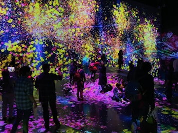 A vibrant and immersive digital art installation with bright, colorful light projections resembling flowers and abstract patterns covering the walls and floor. People are interacting with the environment, some sitting and others standing or walking around, creating an interactive and dynamic atmosphere.