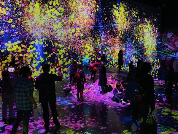 A vibrant and immersive digital art installation with bright, colorful light projections resembling flowers and abstract patterns covering the walls and floor. People are interacting with the environment, some sitting and others standing or walking around, creating an interactive and dynamic atmosphere.