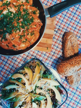 A skillet with cooked shrimp covered in a red sauce and garnished with chopped parsley sits on a wooden board. Nearby, a glass dish contains a salad with avocado slices, seeds, and mixed greens. Two multigrain bread rolls are placed to the side on a checkered tablecloth.