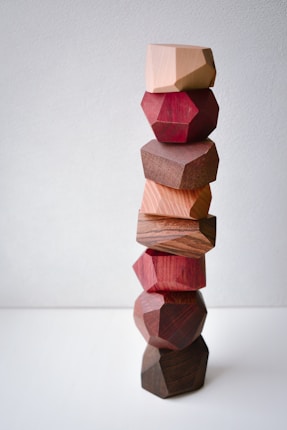 A stack of geometrically shaped wooden blocks in various shades, including natural wood tones and reddish hues, arranged in a stable yet precarious manner against a plain light background.