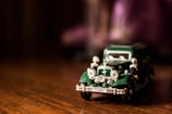 A small, detailed model of a vintage car with a dark green body and chrome accents is placed on a wooden surface. The car has a classic design with rounded headlights and a license plate.