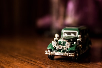 A small, detailed model of a vintage car with a dark green body and chrome accents is placed on a wooden surface. The car has a classic design with rounded headlights and a license plate.