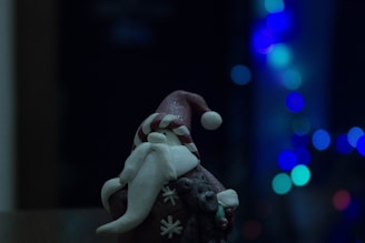 A small figurine of Santa Claus is seen with a long, curled white beard and a red hat with white details. The figure holds a teddy bear and is positioned against a backdrop of blurred blue bokeh lights, creating a festive and cozy atmosphere.
