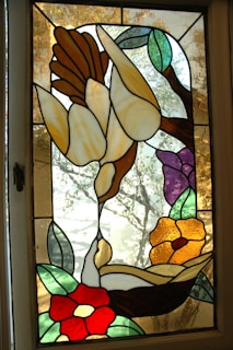 A stained glass window depicting various floral elements, including white flowers with yellowish petals, green leaves, and a red flower with white center. The background has a textured, frosted design with a tree silhouette.