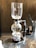 A coffee siphon apparatus is set up on a countertop. It features a glass chamber on top, connected to a spherical bottom chamber. A visible flame is underneath, heating the lower chamber. The setup is sophisticated and suggests precision in brewing coffee.