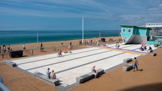A seaside promenade features an inverted house as an unusual attraction. People are walking and sitting on benches along the open, paved area that is directly adjacent to a sandy beach. The sea extends to the horizon under a mostly clear sky. The scene has a mix of tourists observing the novelty house, some strolling along the beach, and others enjoying the sunny weather on the benches.