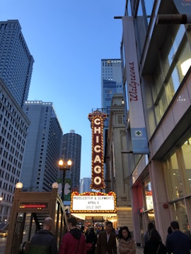 A bustling city street scene with the iconic Chicago Theatre marquee brightly lit. People walk along the sidewalk as skyscrapers tower overhead. The sky is clear and blue, and a Walgreens storefront is visible to the right.