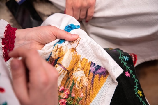 A person is embroidering a colorful image onto fabric, with close detail on the handwork. The scene appears vibrant with intricate patterns being stitched. The person's hands are in focus, working delicately with the needle and thread.