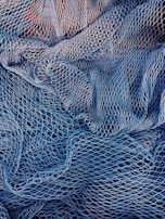 A complex mesh of intertwined fishing nets in varying shades of blue and white. The nets are intricately layered, creating a textured pattern with gaps and openings of different sizes.