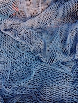 A complex mesh of intertwined fishing nets in varying shades of blue and white. The nets are intricately layered, creating a textured pattern with gaps and openings of different sizes.