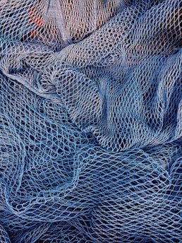 A complex mesh of intertwined fishing nets in varying shades of blue and white. The nets are intricately layered, creating a textured pattern with gaps and openings of different sizes.