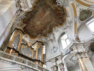 A magnificent baroque-style church interior featuring an ornately decorated pipe organ with golden accents. The ceiling is adorned with intricate frescoes and elaborate ornamental stucco work. Large arched windows allow natural light to illuminate the space, highlighting the rich details of the architecture and the soft pastel color scheme.
