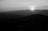 A black and white photograph captures a serene landscape with mountains in the distance. The sun is low on the horizon, creating a gentle glow that outlines the mountain ridges. The foreground features dark silhouettes of rolling hills and rocky terrain.