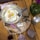 A breakfast setting featuring a pan with three sunny-side-up eggs, topped with black pepper. Beside it, there are slices of dark bread, cheese pieces with black seeds, and chocolate-covered sweets sprinkled with sesame. A green glass vase with pink flowers and a decorative sugar bowl are also present. Two beverages, one in a cup and the other in a glass, complete the setting.