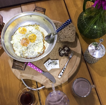 A breakfast setting featuring a pan with three sunny-side-up eggs, topped with black pepper. Beside it, there are slices of dark bread, cheese pieces with black seeds, and chocolate-covered sweets sprinkled with sesame. A green glass vase with pink flowers and a decorative sugar bowl are also present. Two beverages, one in a cup and the other in a glass, complete the setting.