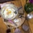 A breakfast setting featuring a pan with three sunny-side-up eggs, topped with black pepper. Beside it, there are slices of dark bread, cheese pieces with black seeds, and chocolate-covered sweets sprinkled with sesame. A green glass vase with pink flowers and a decorative sugar bowl are also present. Two beverages, one in a cup and the other in a glass, complete the setting.