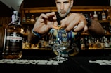 A person is preparing a drink at a bar, with a focus on their hands skillfully twisting an orange peel over a glass. In the foreground, there is a bottle labeled Drambuie. The background features shelves filled with various liquor bottles, suggesting a bar setting. The lighting is warm, enhancing the cozy atmosphere.