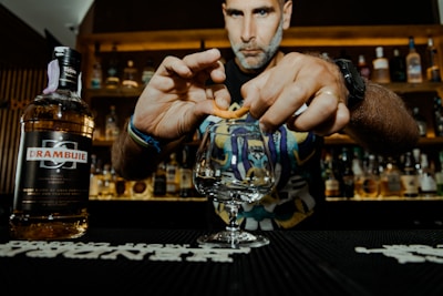 A person is preparing a drink at a bar, with a focus on their hands skillfully twisting an orange peel over a glass. In the foreground, there is a bottle labeled Drambuie. The background features shelves filled with various liquor bottles, suggesting a bar setting. The lighting is warm, enhancing the cozy atmosphere.