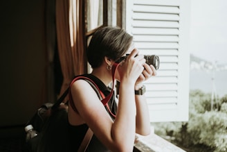A person with short hair stands by an open window, holding a camera up to their face as they take a photograph. They are wearing a sleeveless top and carrying a backpack with a water bottle tucked into a side pocket. Light filters in through the window, illuminating the person's profile, while the scenery outside includes greenery and a distant view.