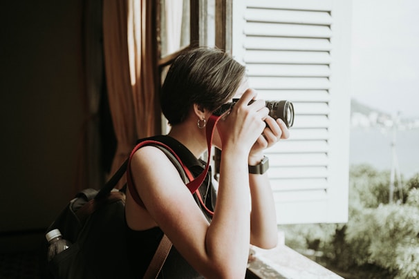 A person with short hair stands by an open window, holding a camera up to their face as they take a photograph. They are wearing a sleeveless top and carrying a backpack with a water bottle tucked into a side pocket. Light filters in through the window, illuminating the person's profile, while the scenery outside includes greenery and a distant view.