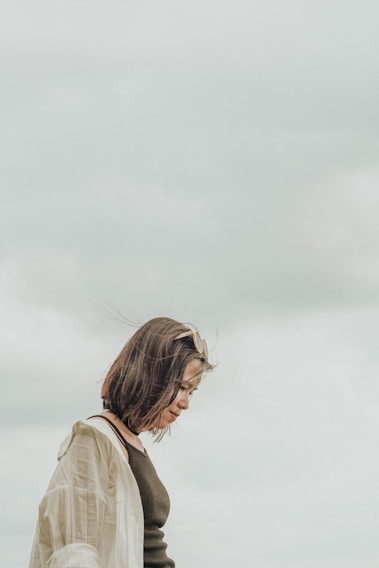 A person with short brown hair looks downward, wearing sunglasses and a light jacket against a cloudy sky backdrop. The composition conveys a sense of contemplation or introspection.