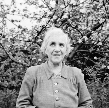 An elderly woman with short, light-colored hair is standing in front of a blossoming tree. She is wearing a buttoned-up blouse with a decorative brooch and appears to be smiling gently. The background is filled with branches and small flowers, creating a natural and serene setting.
