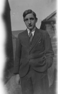 A young person stands outdoors, dressed in a formal suit with a tie. The setting appears to be an alley or narrow passage, with brick buildings and a clear sky in the background. The pose is relaxed with one hand in the pocket.