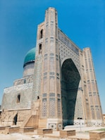 A grand historical building with intricate mosaic patterns and geometric designs covers the facade. The structure features a large archway and a prominent turquoise dome, characteristic of Islamic architecture. The brickwork and decorative tiles exhibit a blend of earthy and vibrant colors, reflecting meticulous craftsmanship.