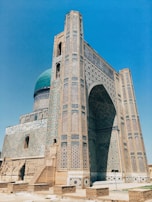 A grand historical building with intricate mosaic patterns and geometric designs covers the facade. The structure features a large archway and a prominent turquoise dome, characteristic of Islamic architecture. The brickwork and decorative tiles exhibit a blend of earthy and vibrant colors, reflecting meticulous craftsmanship.